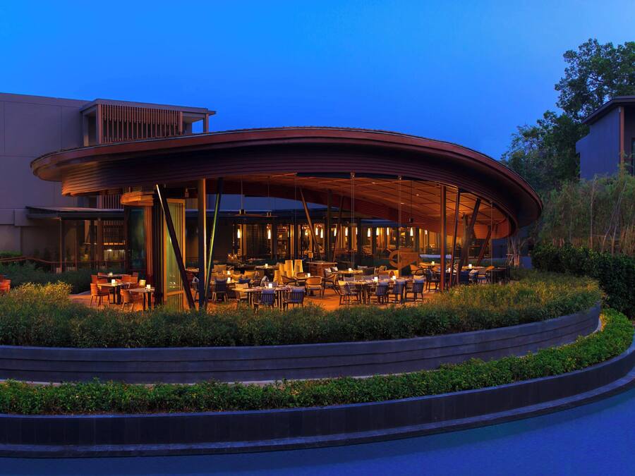 A modern outdoor dining area with a curved roof, surrounded by greenery and a pool.