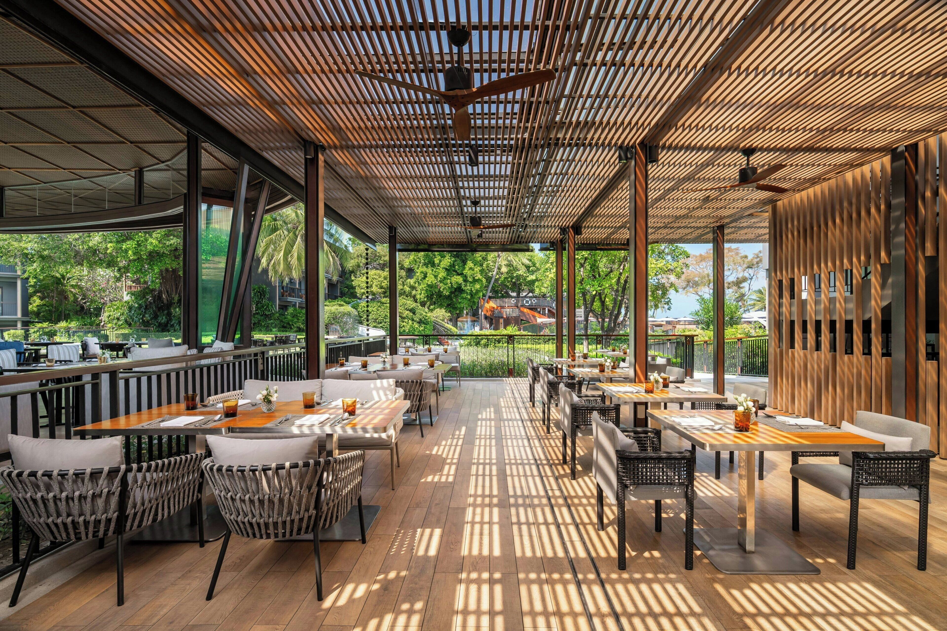 An outdoor dining area with wooden tables and chairs, large windows, and a view of greenery.