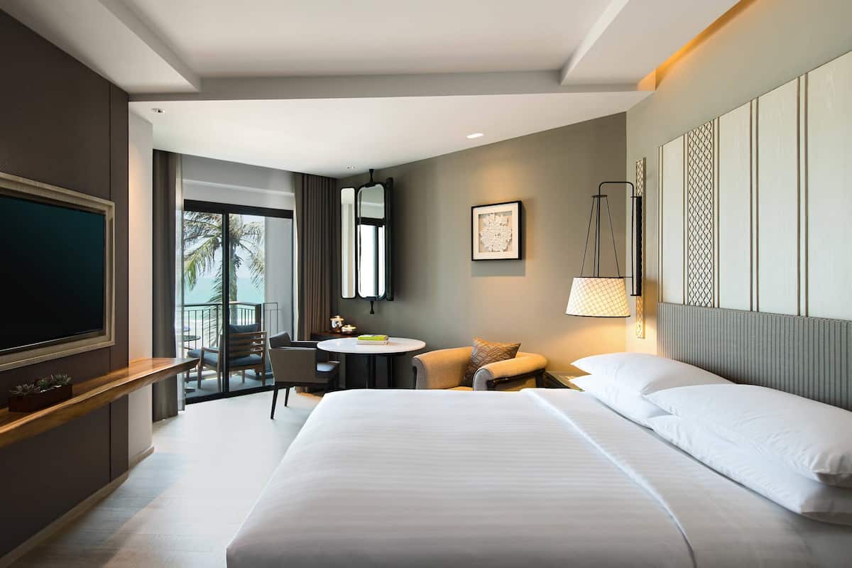 A hotel room with a large bed, a TV, a desk with a chair, a small table, and a balcony with a view of palm trees.