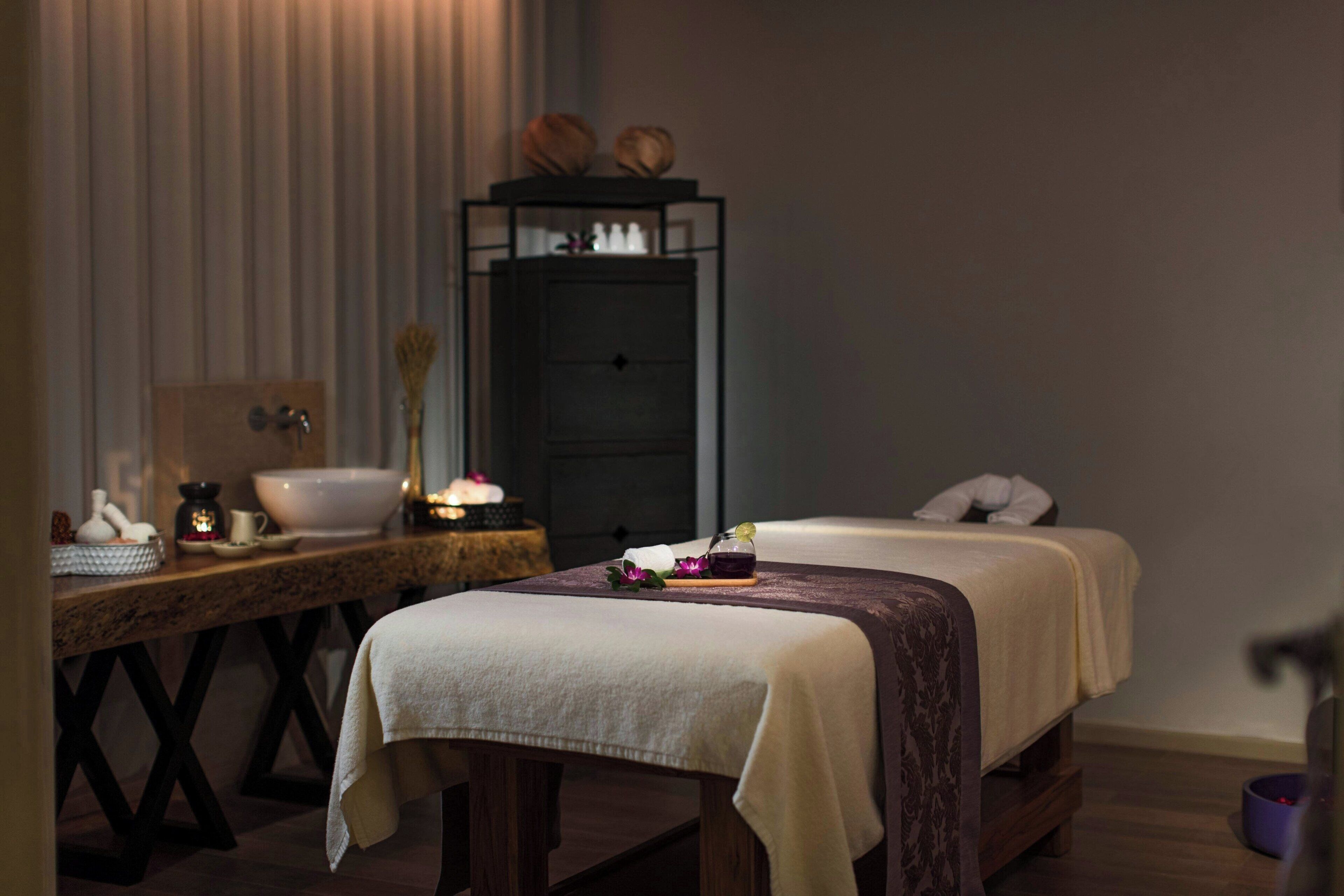 A spa room with two beds, a wooden table with a bowl and candles, and a dark cabinet with decorative items.