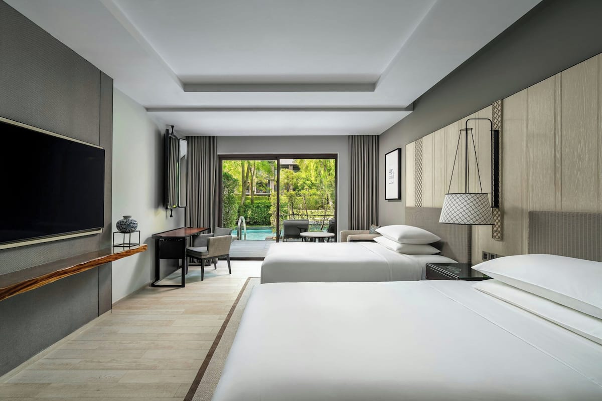 Een moderne hotelkamer met een groot bed, een bureau, een stoel, een televisie en uitzicht op een zwembad en groen.