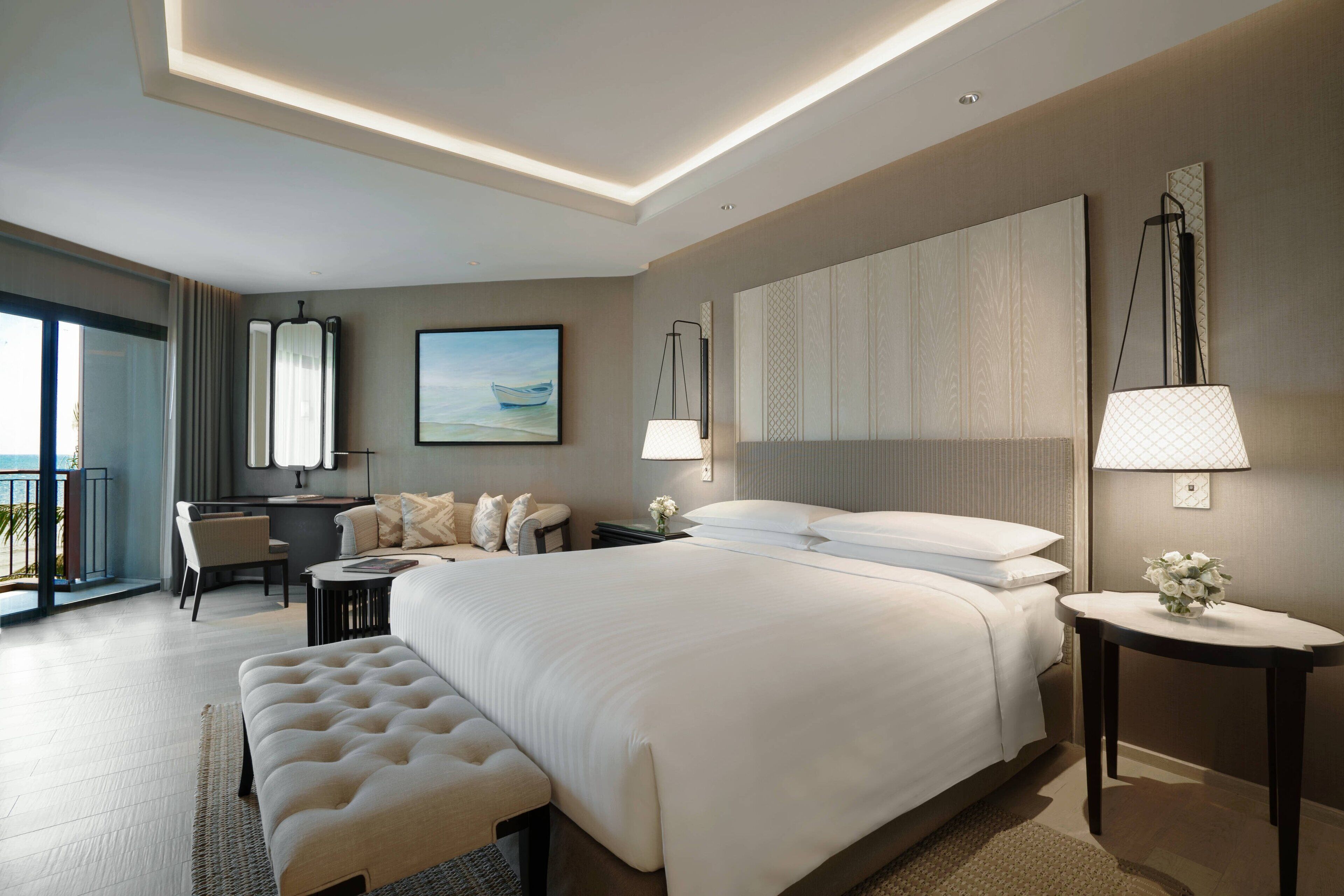 A modern hotel room with a large bed, a sitting area with a sofa and armchairs, a desk, and a view of the outdoors.