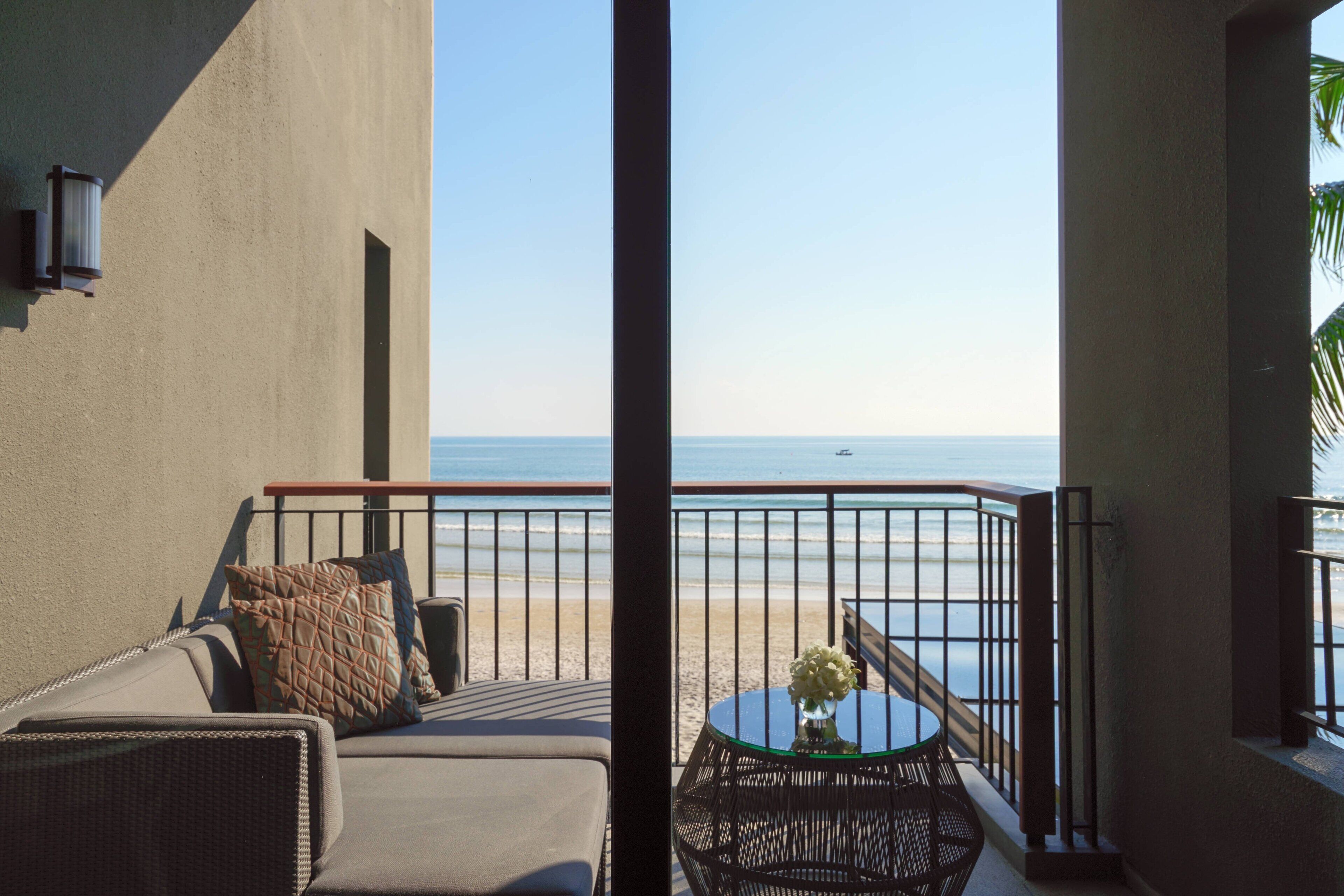 A balcony with a view of the beach, a round table with a vase of flowers, and a sofa with cushions.