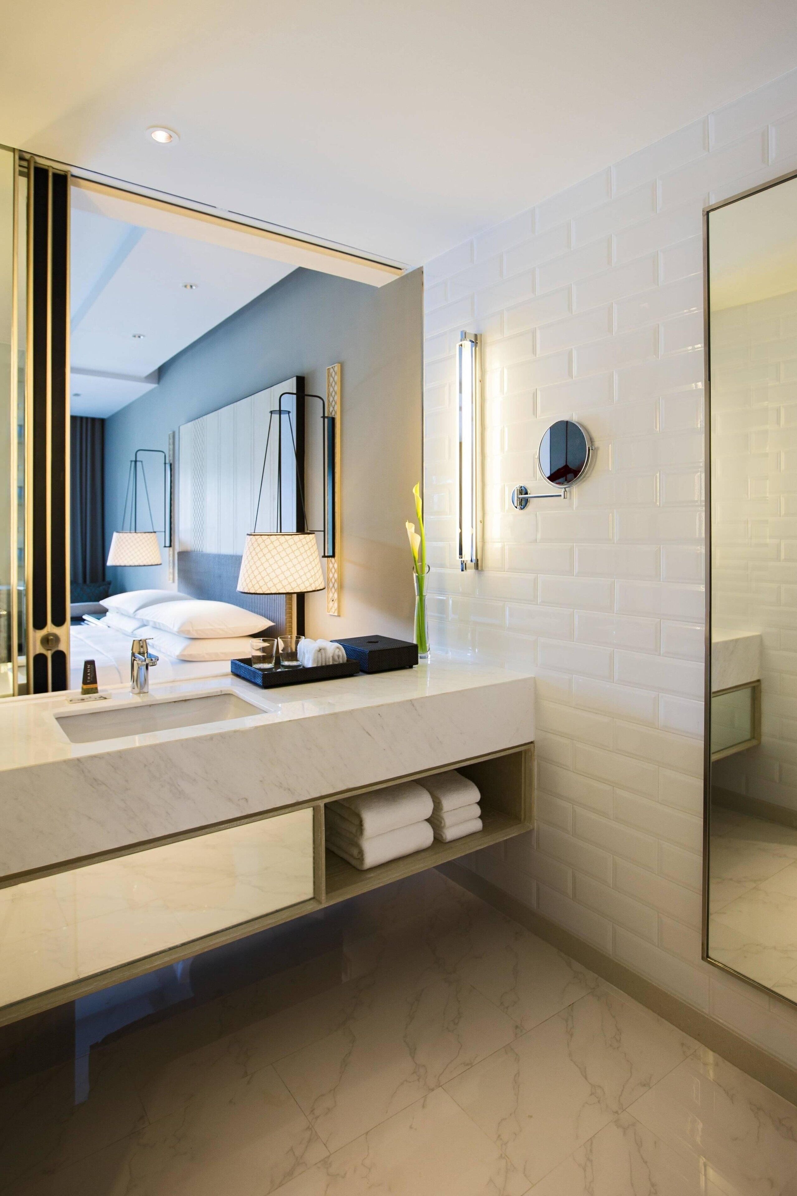 Un baño moderno con un espejo grande, paredes de azulejos blancos, una encimera de mármol y una cama visible al fondo.