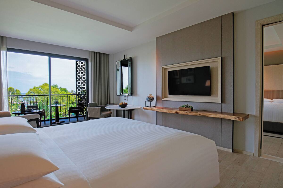 Een hotelkamer met een groot bed, een aan de muur bevestigde televisie, een klein tafeltje met een vaas en een balkon met uitzicht op bomen.