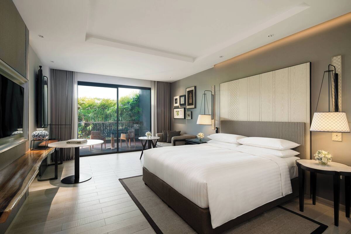 Een moderne hotelkamer met een groot bed, een zithoek met een tafel en stoelen, een balkon met uitzicht op groen, en een groot raam.