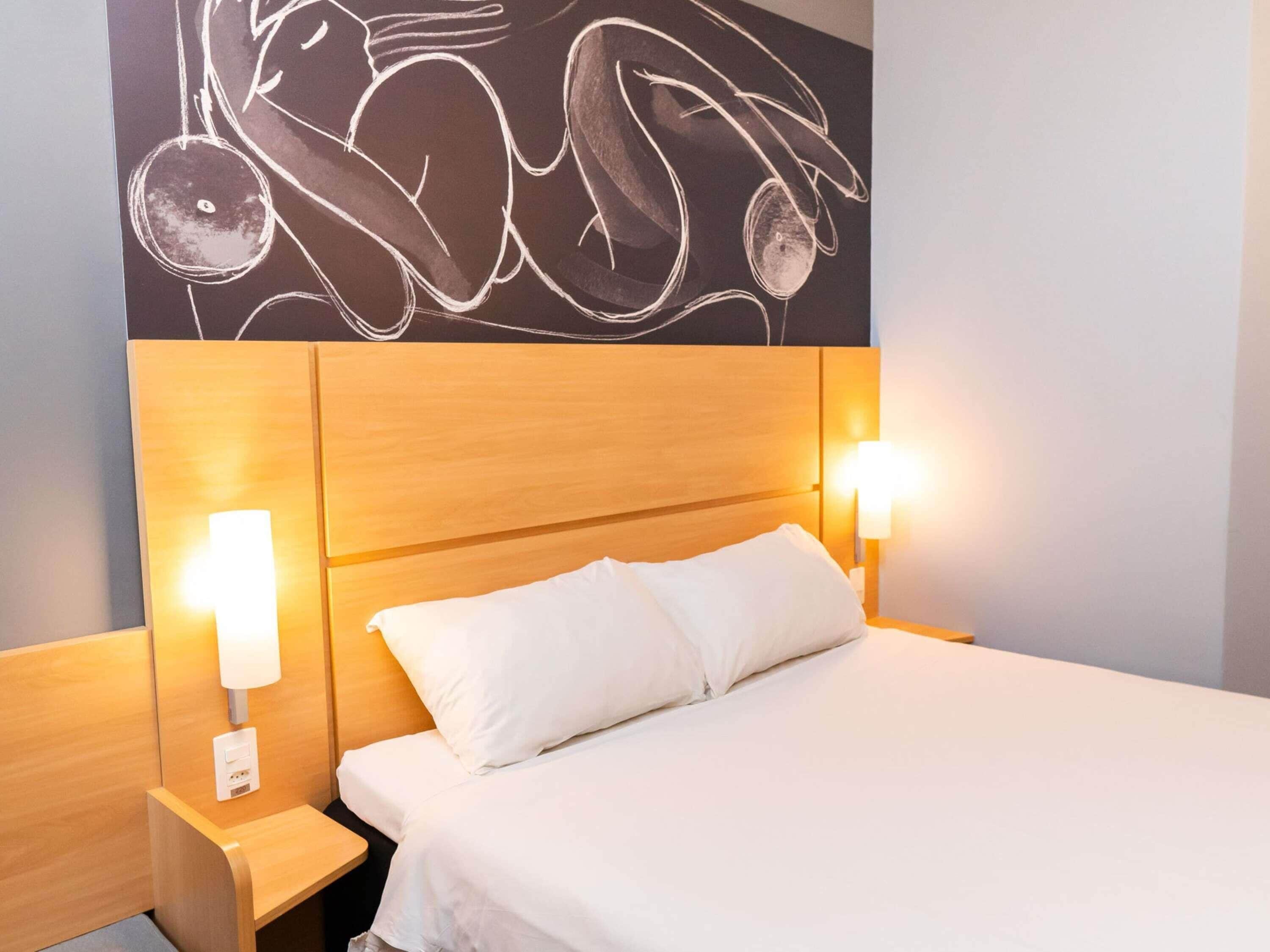 A hotel room with a wooden headboard, two bedside lamps, a white bed, and a wall with abstract art.