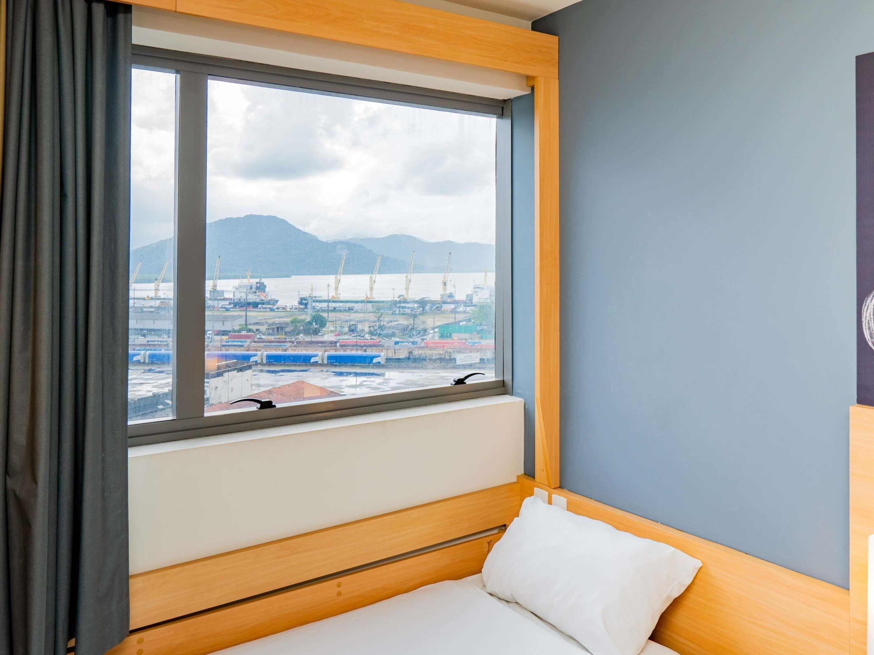 A hotel room with a large window, a bed, and a view of a port with ships and cranes.