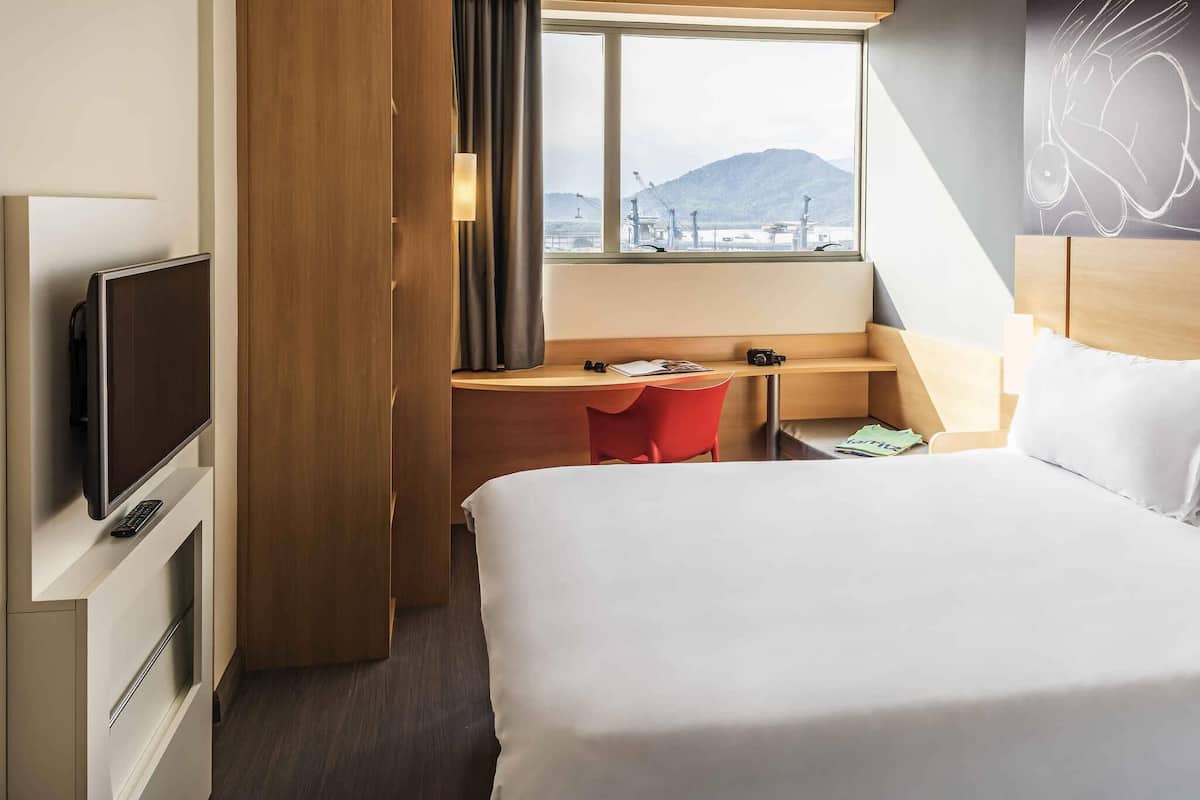 A hotel room with a bed, a desk, a chair, a television, and a window with a view of a mountain and industrial area.