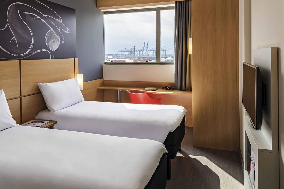 A hotel room with two beds, a desk, a TV, and a window with a view of a port.