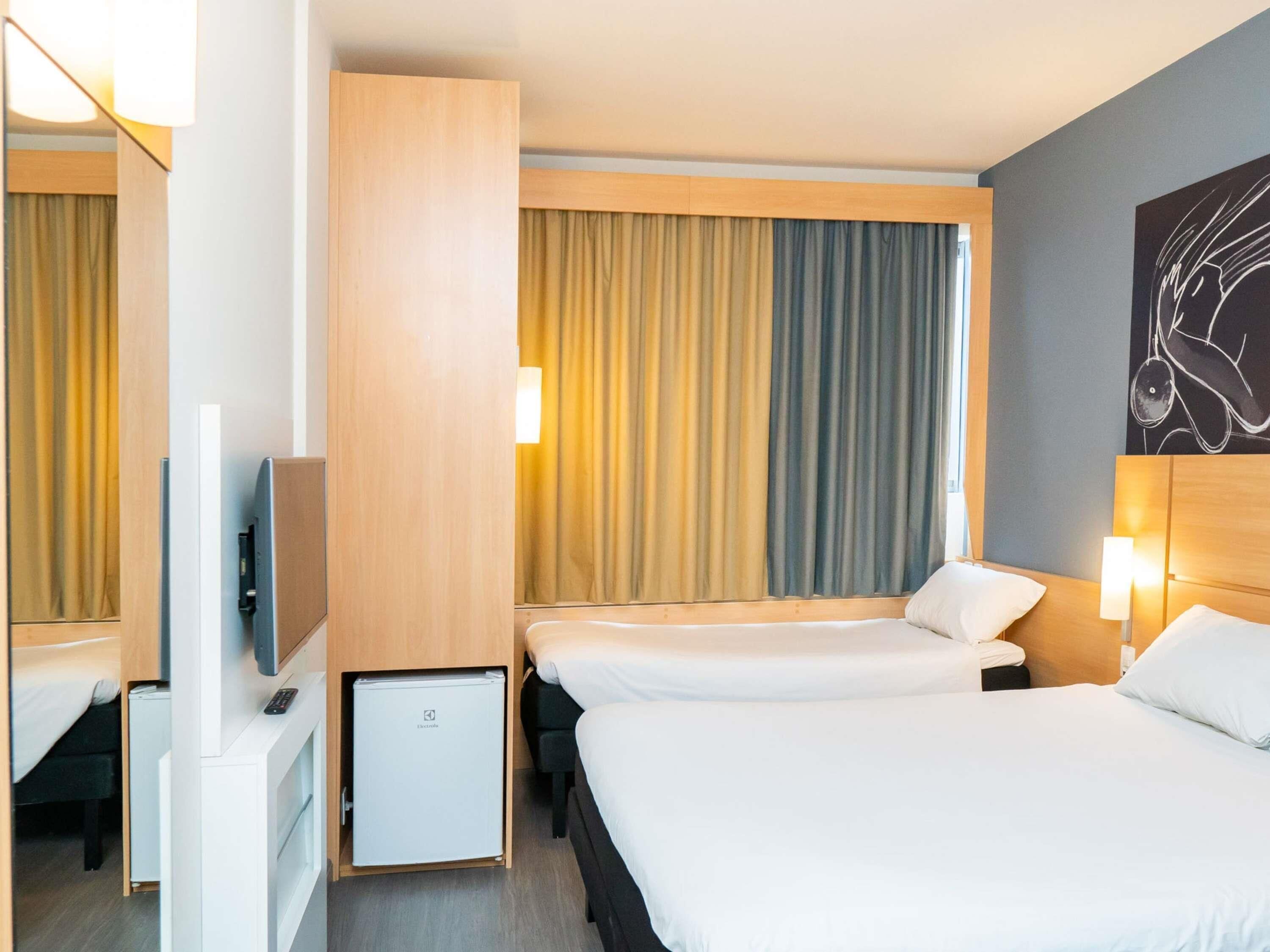 Ein Hotelzimmer mit zwei Betten, einem Fernseher, einer Minibar und einem großen Fenster mit Vorhängen.