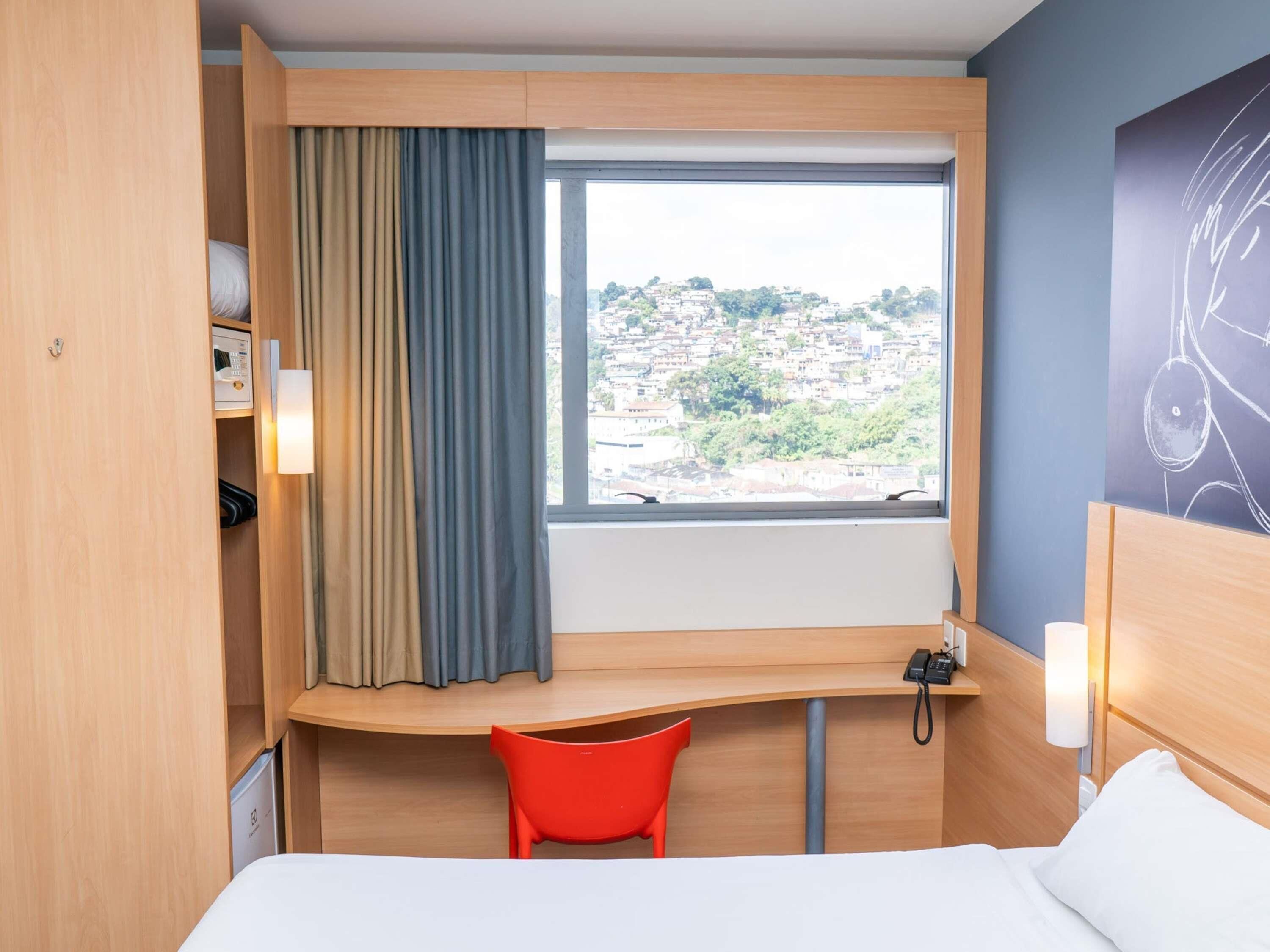 A hotel room with a bed, a desk, a chair, a window with curtains, and a view of a cityscape.