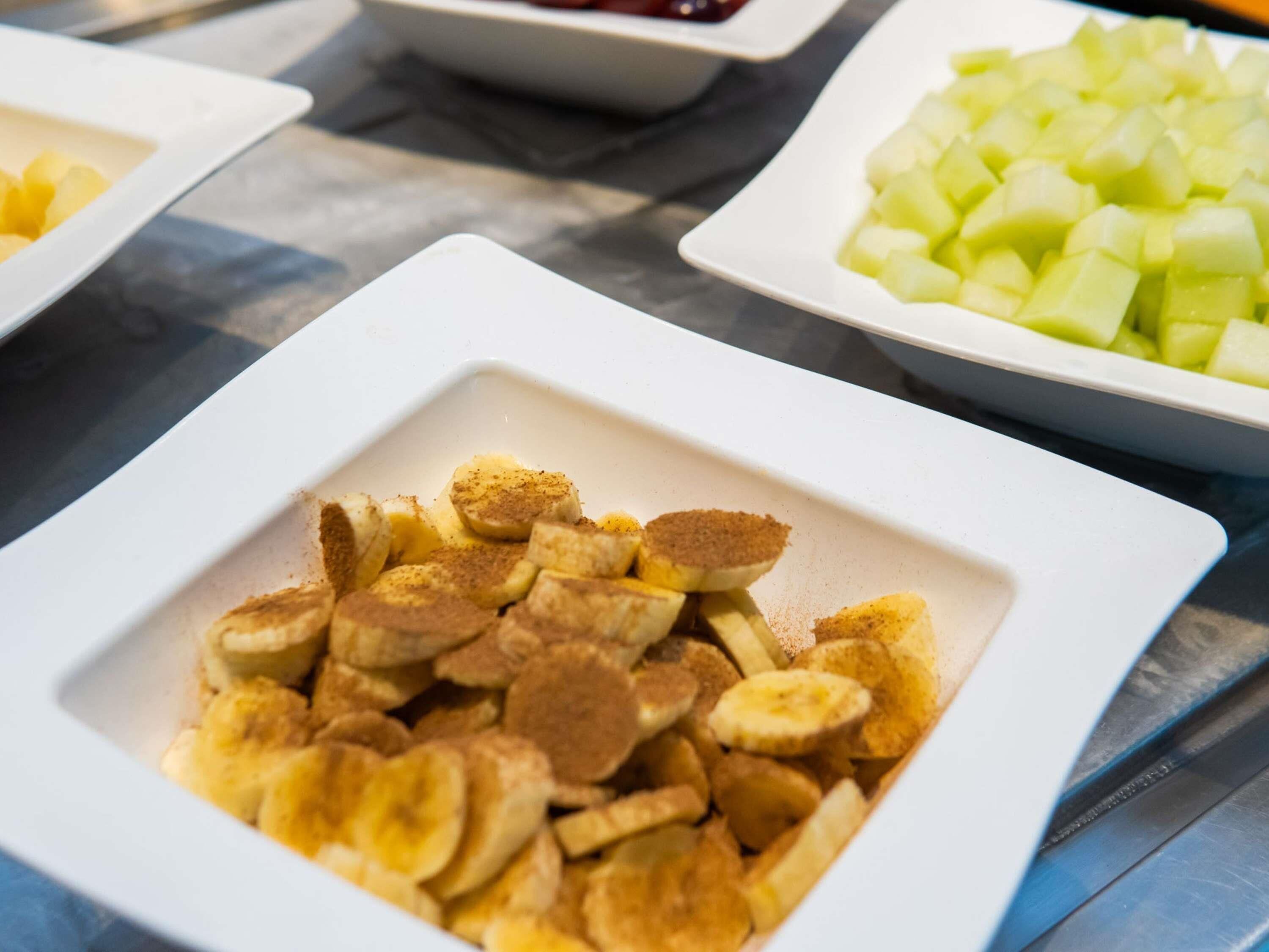 A table with square white plates containing sliced bananas and a white bowl with diced fruit.