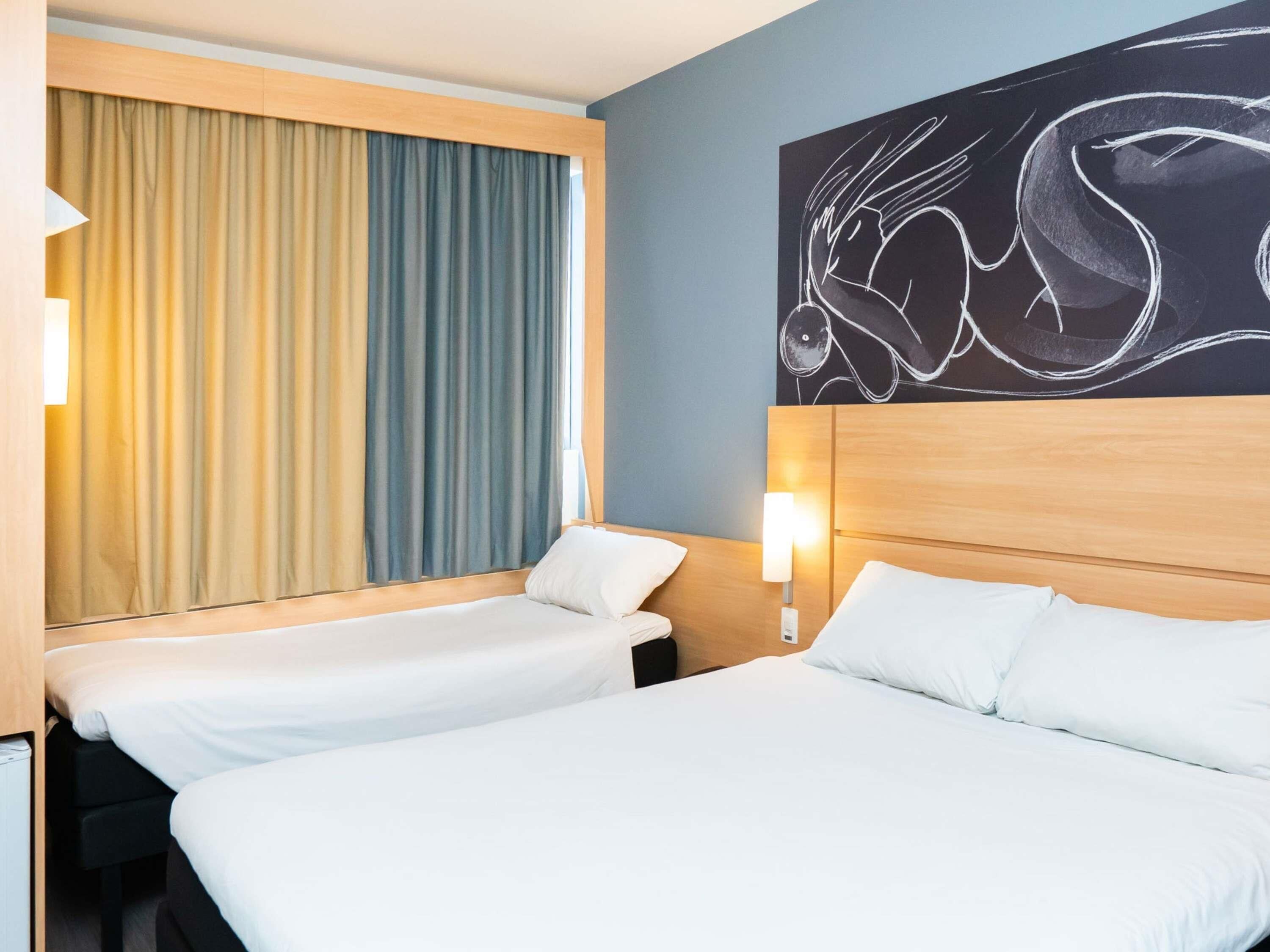 A hotel room with two beds, a large window with curtains, a wall art piece, and a bedside lamp.