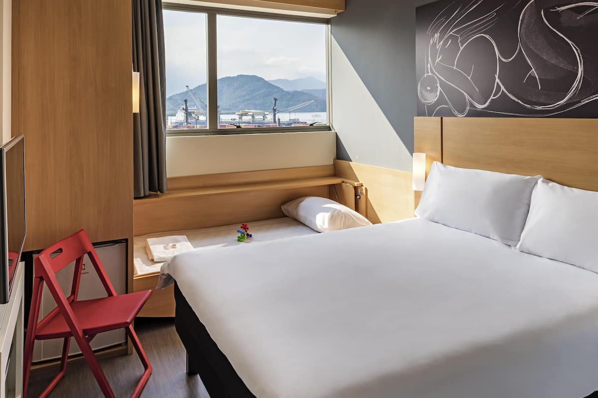 A hotel room with a bed, a desk, a chair, a window with a view of mountains, and a wall with a drawing.