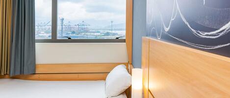 Un lit simple avec une tête de lit en bois, une lampe de chevet et une vue sur un port avec des grues, visible par une fenêtre.