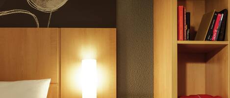 Une chambre d’hôtel avec un lit, une table de chevet sur laquelle se trouvent une tablette et des écouteurs, une lampe fixée au mur et une bibliothèque contenant des livres et un objet rouge.