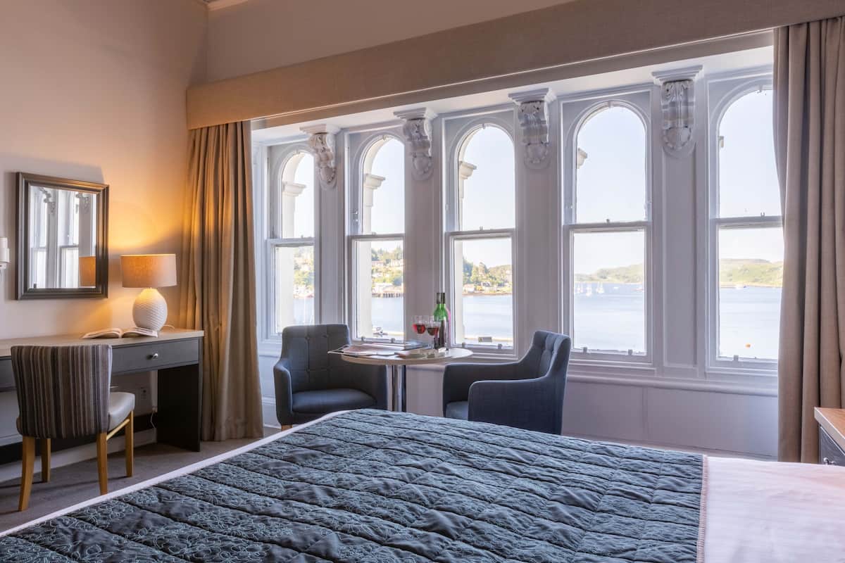 A hotel room with a large bed, a desk with a lamp, two chairs, a small table with a bottle and glasses, and large windows with a view of the water.