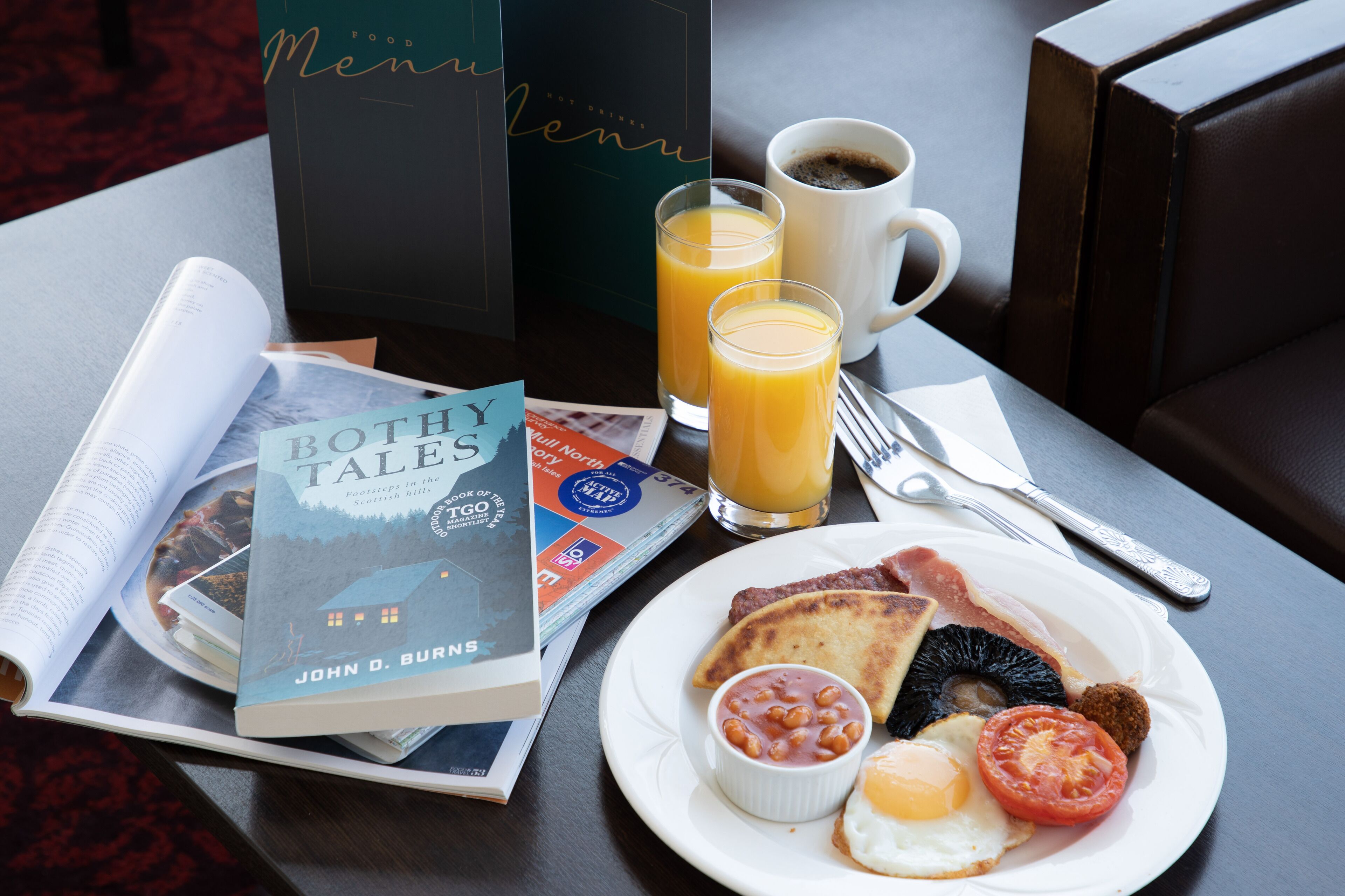 Une table mise pour le petit-déjeuner avec un livre, une assiette d’œufs, de haricots et de pain grillé, une tasse de café et un verre de jus d’orange.