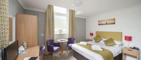 A hotel room with a bed, desk, chairs, a TV, and a window with curtains.