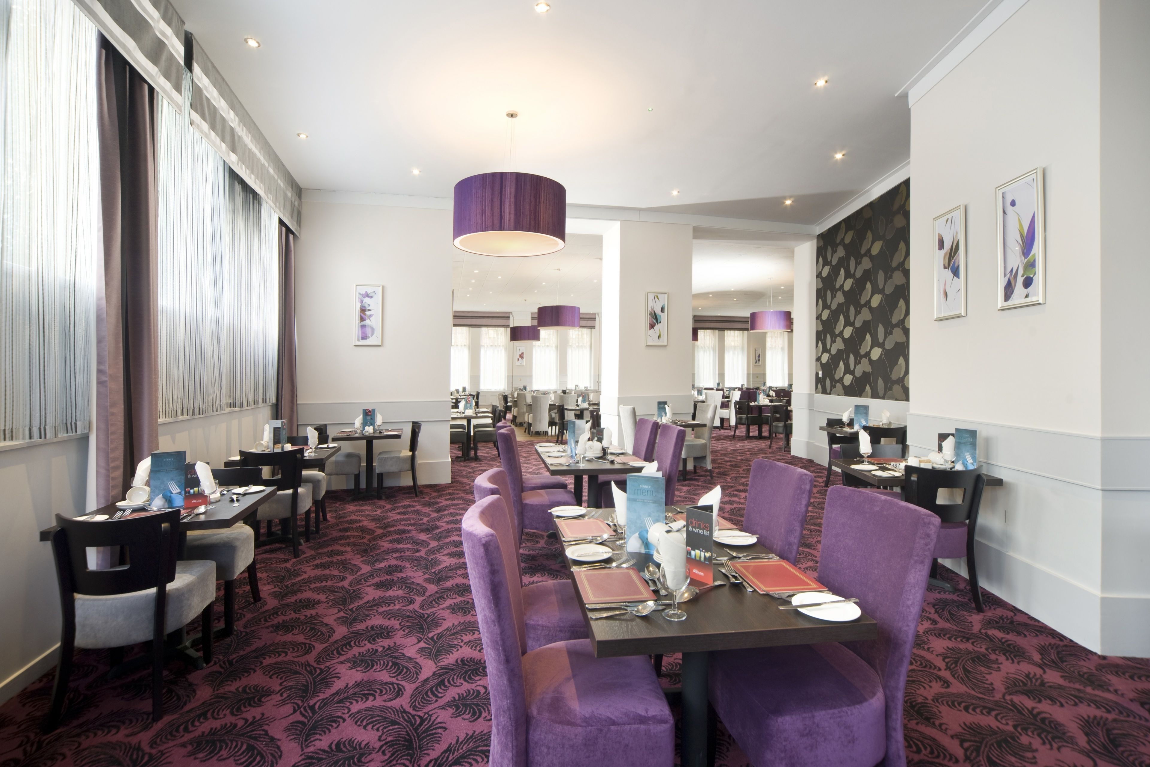 Une salle à manger d’hôtel avec de nombreuses tables dressées, des chaises à revêtement violet et un tapis à motifs.