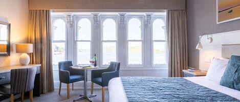 Una habitación de hotel con una cama grande, un escritorio con una silla, una mesita con una lámpara y una ventana con vistas al campo.