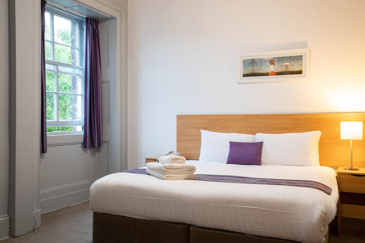 A neatly made bed with a headboard, a bedside table with a lamp, and a window with curtains.