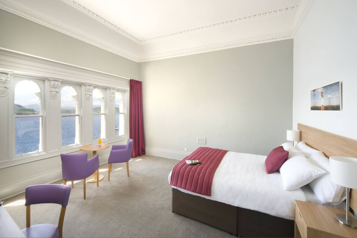 A hotel room with a large bed, a desk with chairs, a view of the sea, and a painting on the wall.