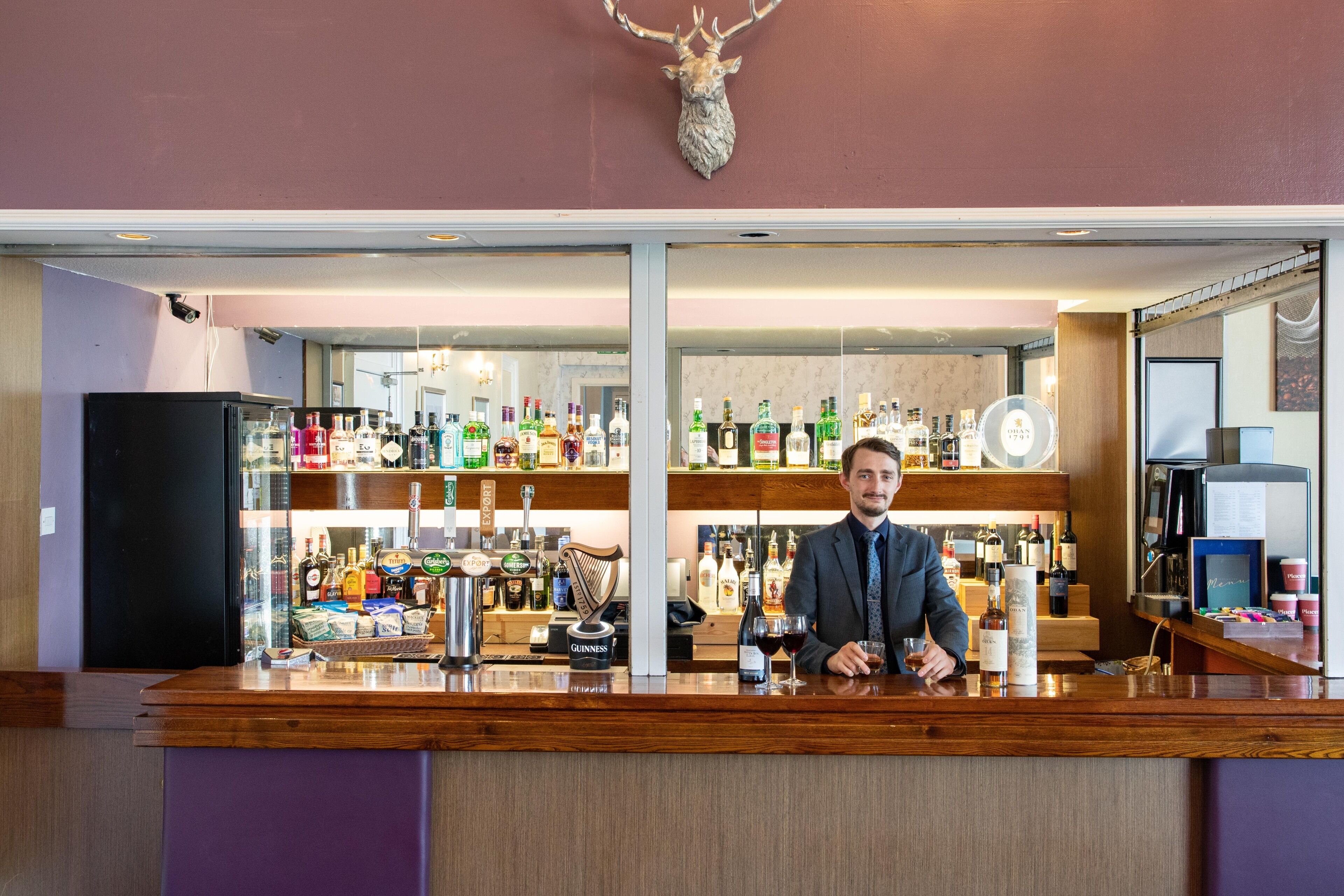 A bar area with a bartender, shelves stocked with various liquor bottles, and a mounted deer head on the wall.