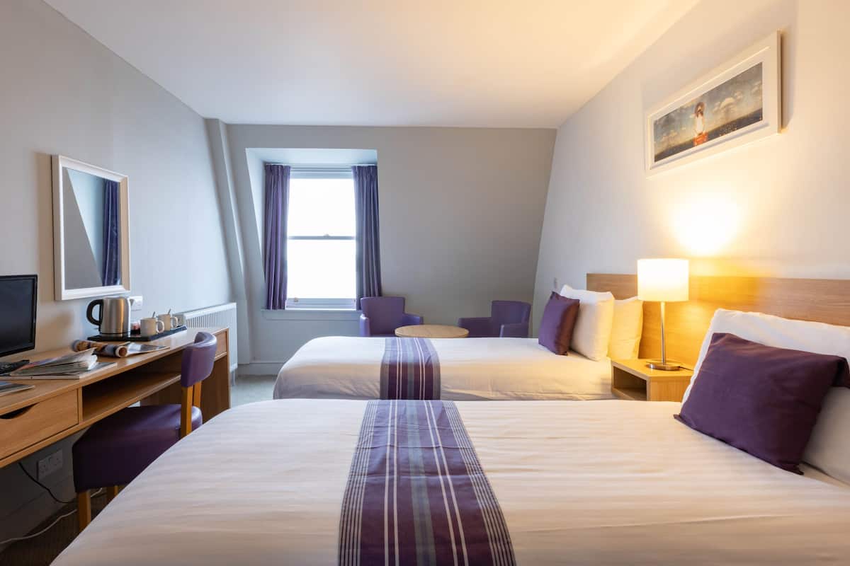 A hotel room with two beds, a desk with a computer, a chair, a lamp, and a window with curtains.