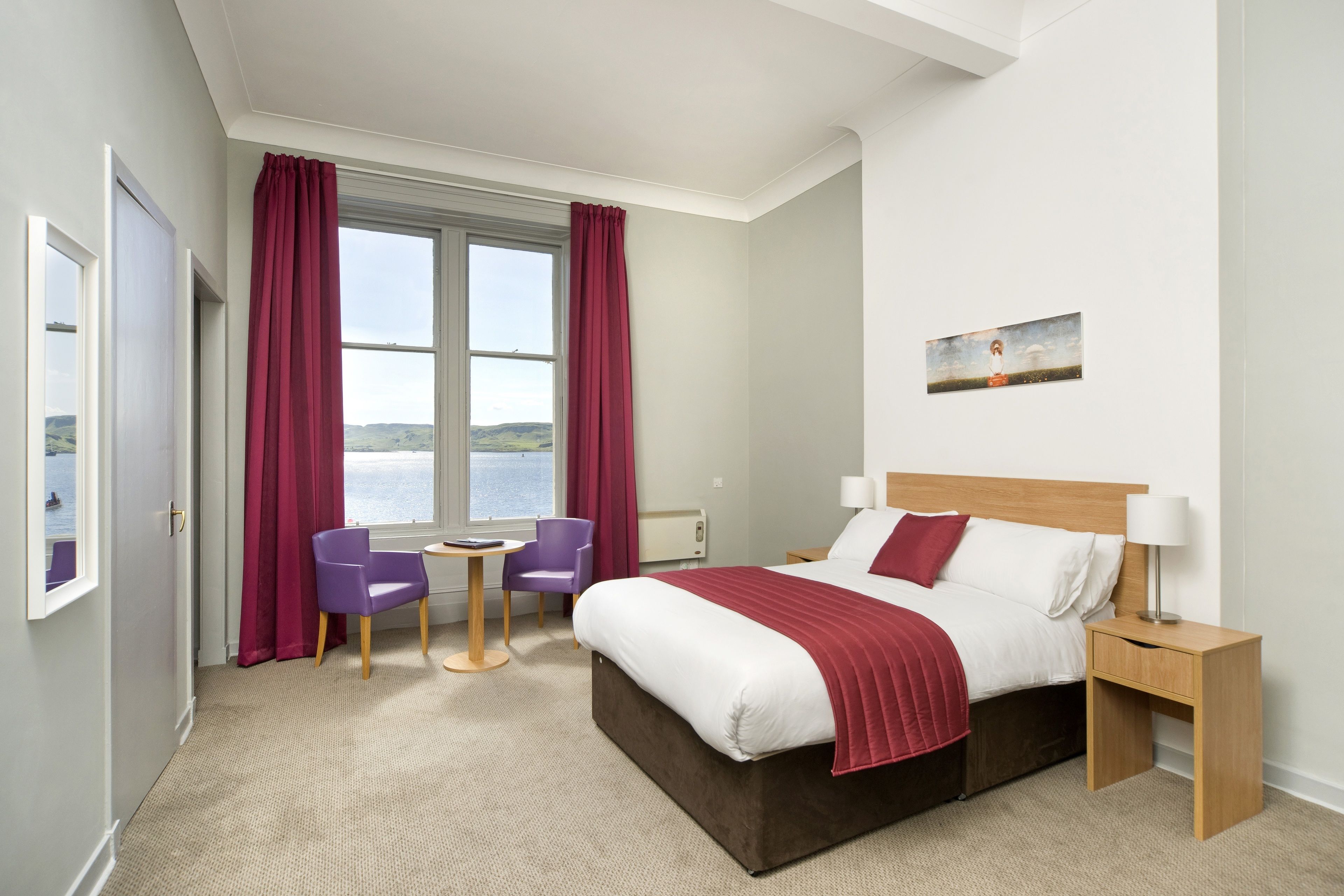 Een hotelkamer met een groot bed, een klein tafeltje, twee stoelen, een nachtkastje en een schilderij aan de muur.