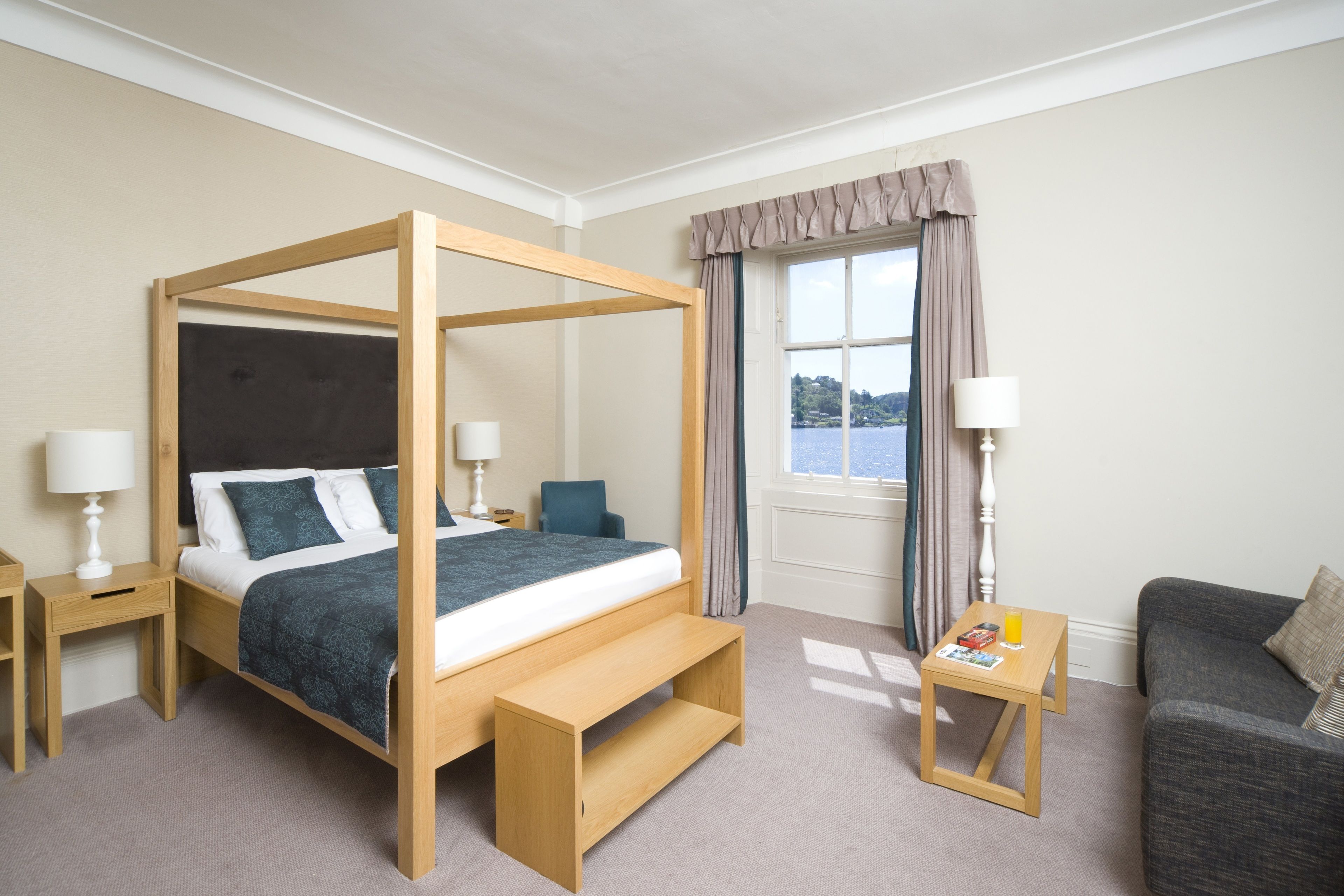 A four-poster bed with a wooden frame and a dark blue bedspread, a wooden nightstand with a lamp, a small wooden table with a lamp, a grey sofa, and a window with curtains.