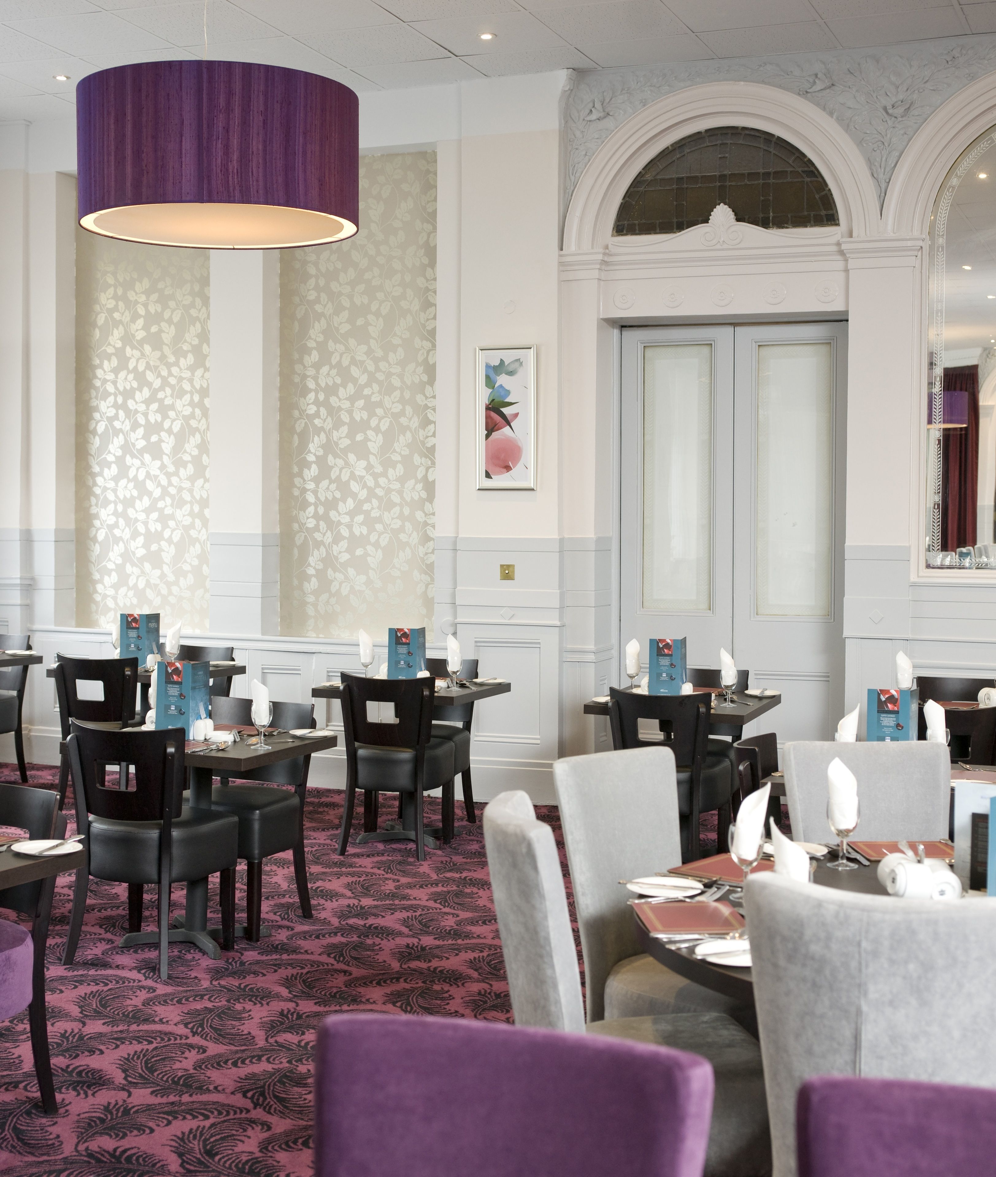 Un restaurant doté d’un luminaire suspendu de couleur violette, d’un papier peint à motifs floraux et d’une grande porte en arc de cercle.