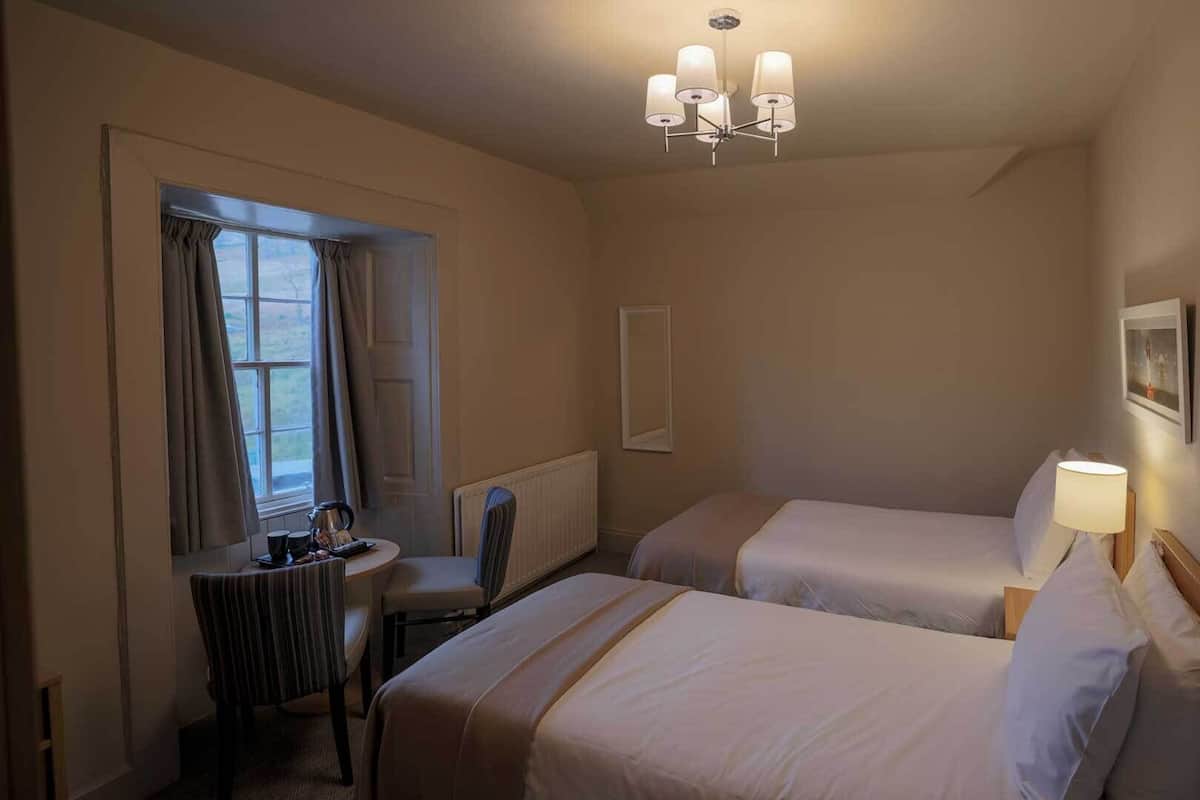 A hotel room with two beds, a small table with a chair, a window with curtains, a radiator, and a chandelier.