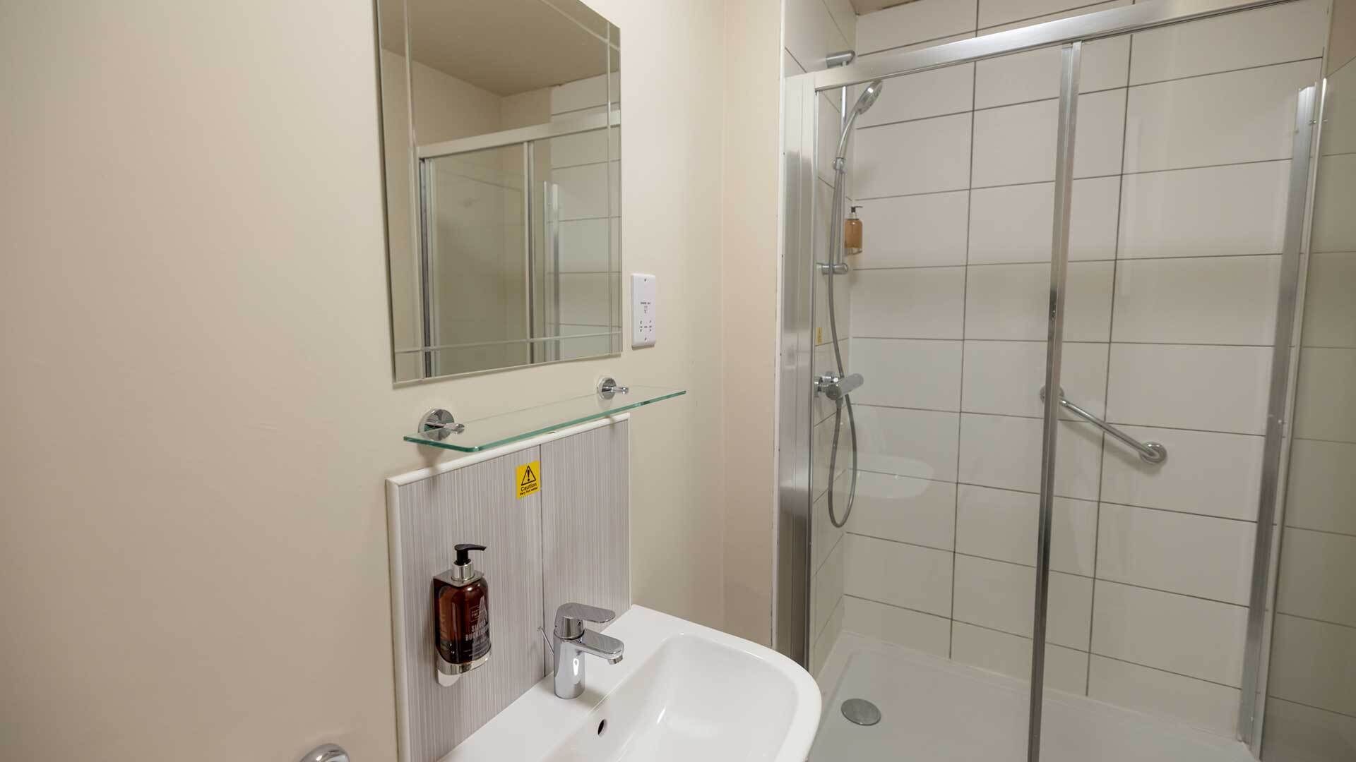 Ein Badezimmer mit Duschkabine aus Glas, Waschbecken mit Spiegel darüber und Seifenspender an der Wand.