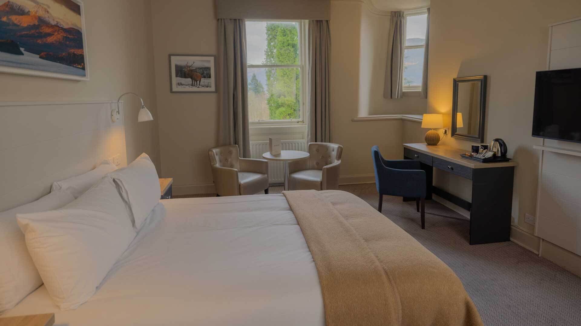 Habitación de hotel con una cama grande, dos sillones, un escritorio, televisión y una ventana con cortinas.