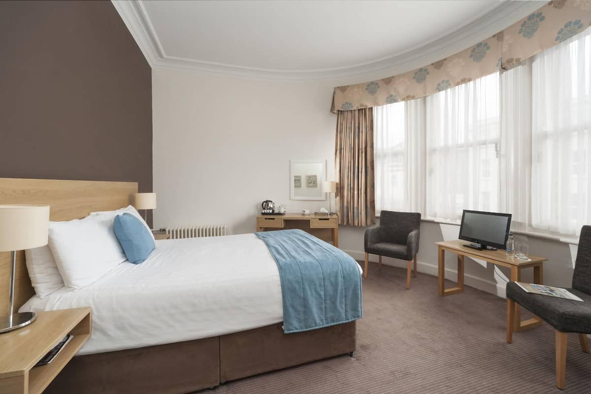A hotel room with a bed, a desk, a chair, a TV, and a window with curtains.