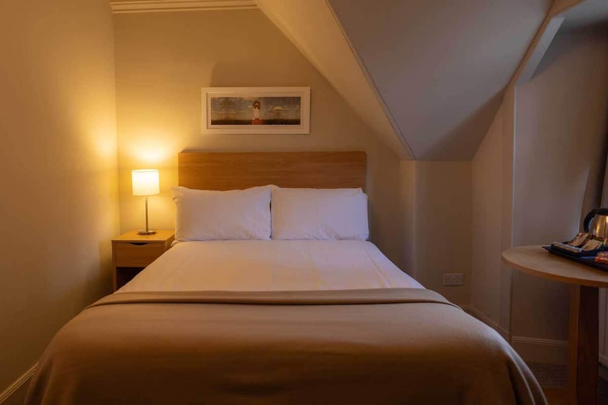 A hotel room with a bed, a nightstand with a lamp, a small table with a kettle, and a painting on the wall.