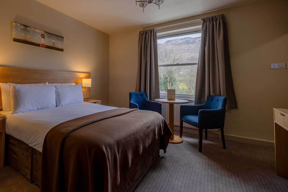 A hotel room with a large bed, two bedside lamps, a small round table, two blue chairs, and a window with curtains.