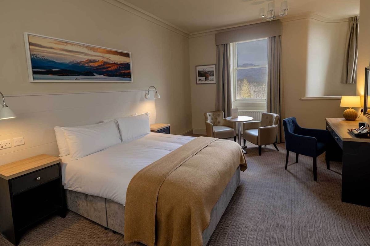 A hotel room with a bed, bedside tables, a desk with a chair, a TV, and a window with a view of mountains.