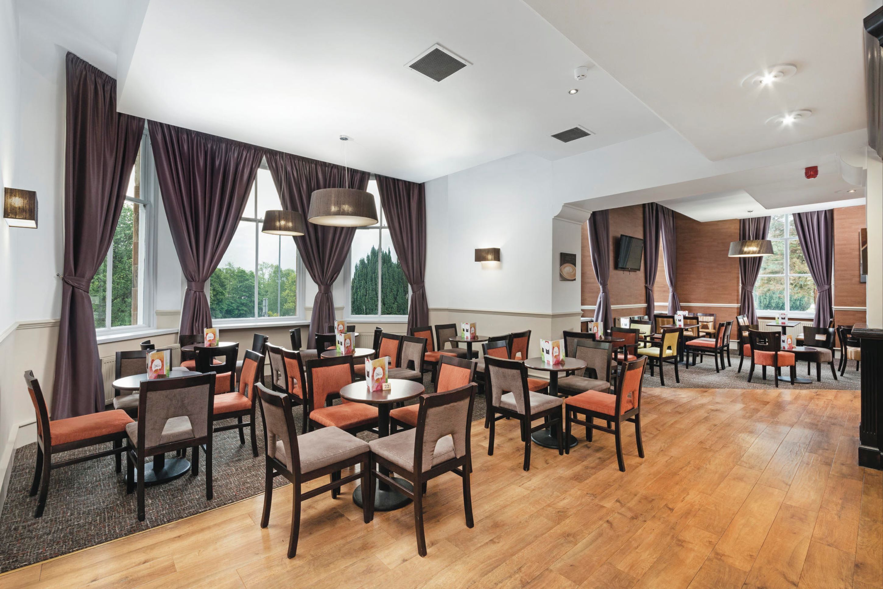 A spacious dining area with multiple tables and chairs, large windows with curtains, and a modern ceiling design.