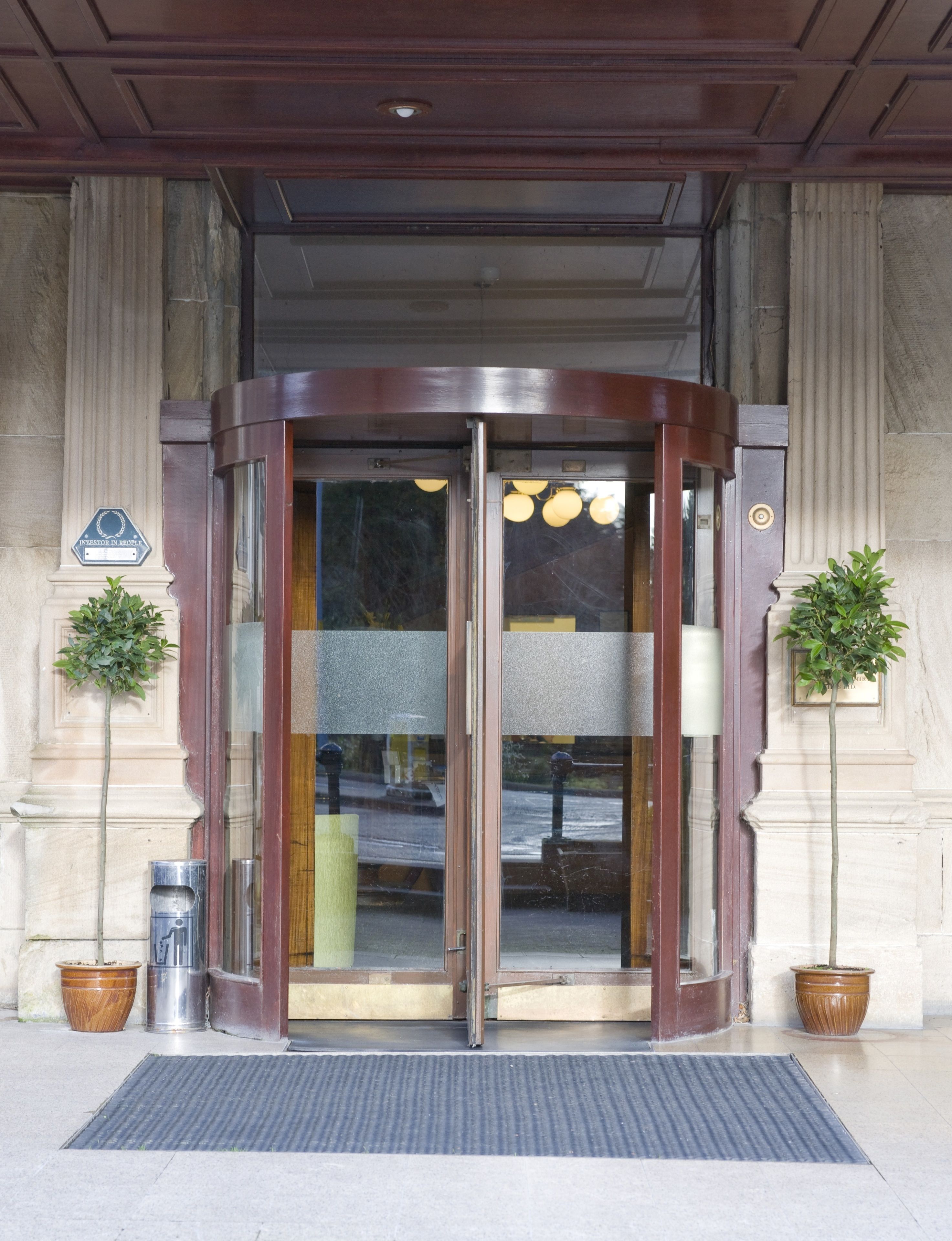 A revolving door with glass panels, flanked by two potted plants, leading to a building entrance with a metal door and a doormat.