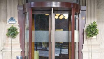 A revolving door with glass panels, flanked by two potted plants, leading to a building entrance with a metal door and a doormat.