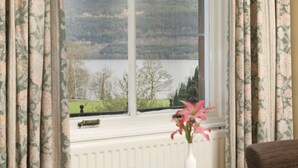 A room with a view of a lake and hills, a round table with a vase of flowers, and floral curtains. - The Tarbet Hotel (Arrochar)
