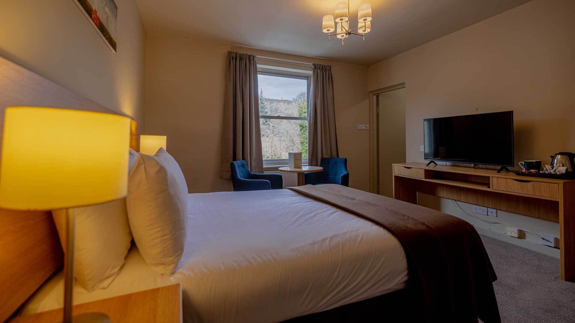 Habitación de hotel con cama, lámpara de noche, televisor, escritorio, silla y ventana con cortinas.