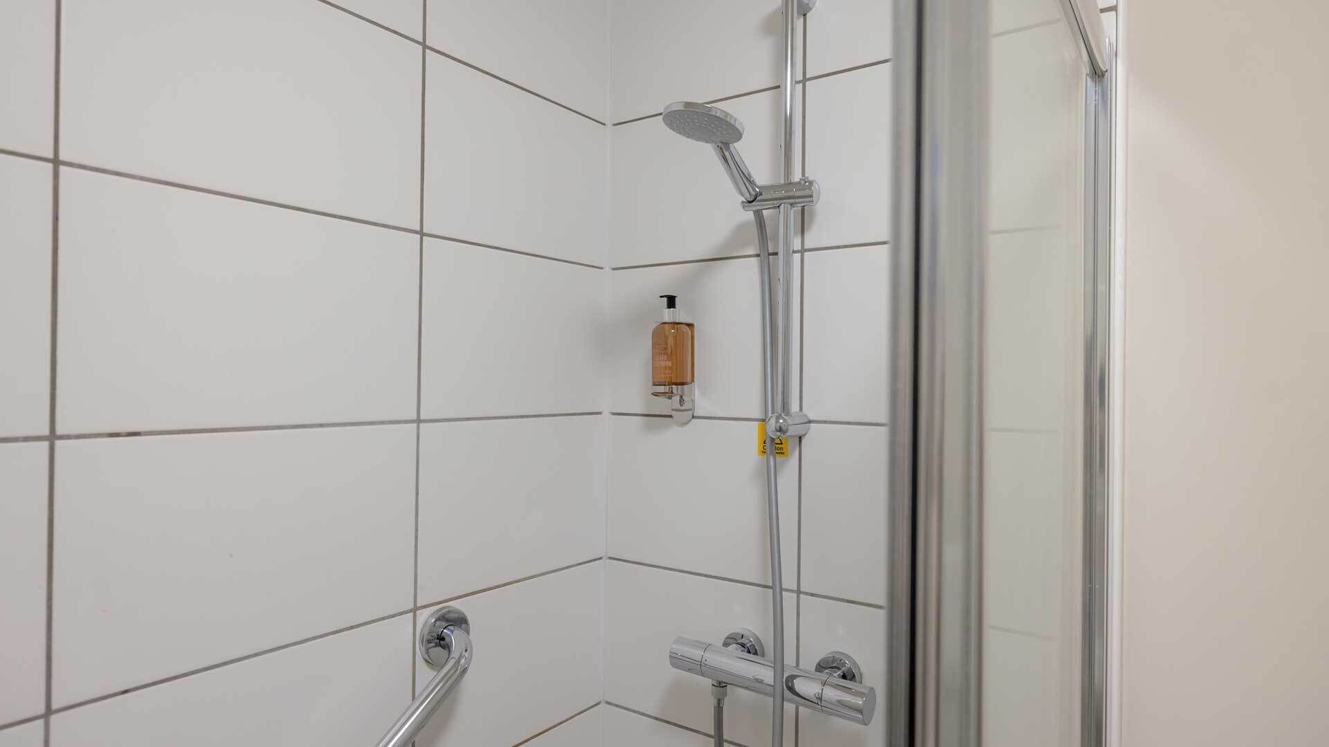 A bathroom with white tiled walls, a glass shower enclosure, a handheld showerhead, a handheld showerhead, and a soap dispenser.