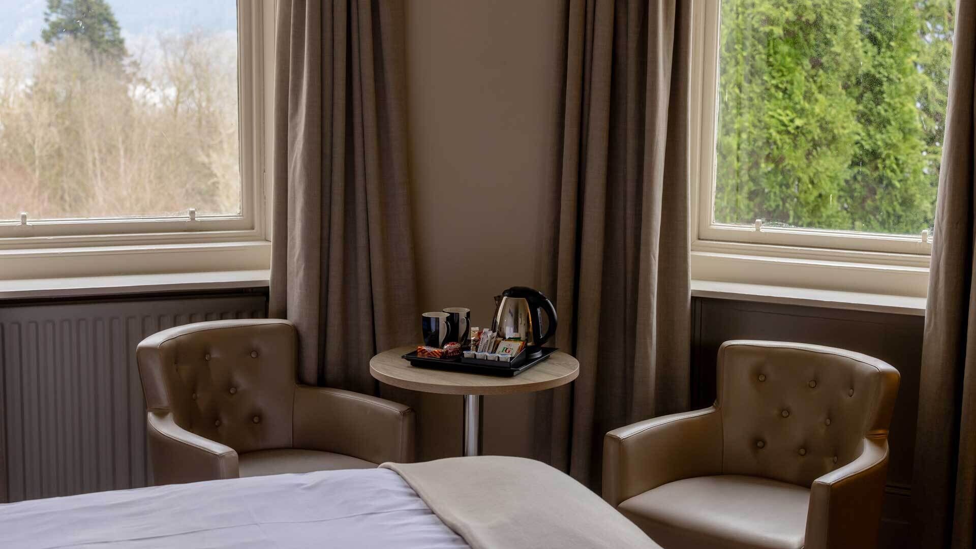 Habitación de hotel con cama, dos sillones, una mesita con cafetera y vistas a árboles por la ventana.