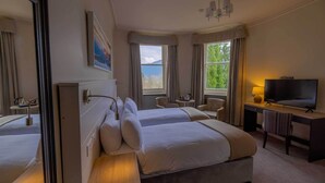 A hotel room with two beds, a television, a desk with a lamp, a chair, and a window with a view of greenery. - The Tarbet Hotel (Arrochar)