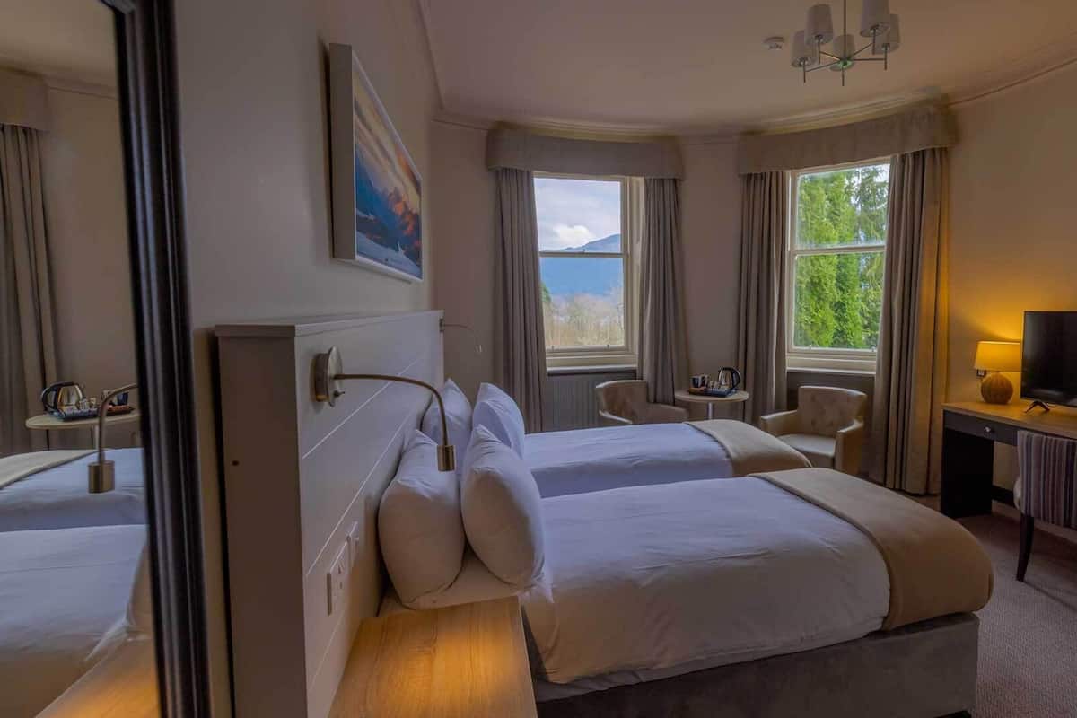 A hotel room with two beds, a television, a desk with a lamp, a chair, and a window with a view of greenery.