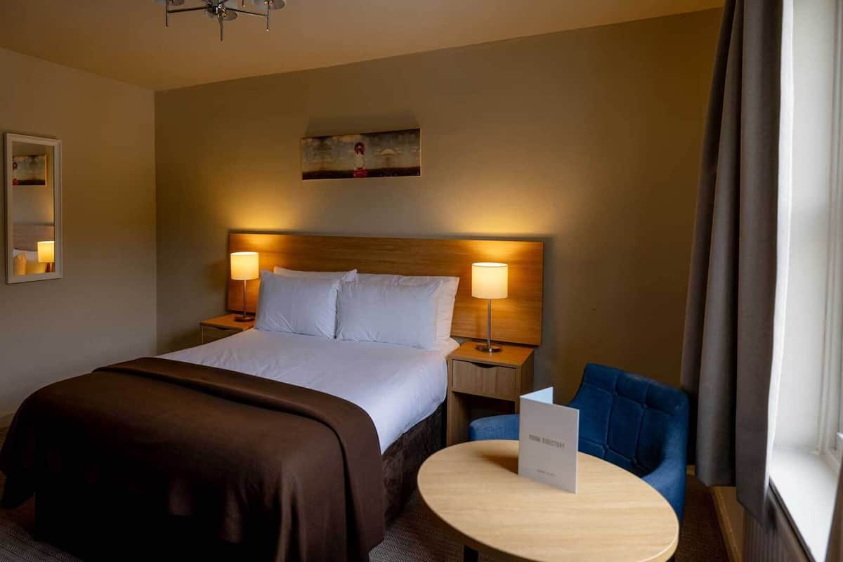 A hotel room with a large bed, two bedside lamps, a small table, a chair, a painting on the wall, and a window with curtains.